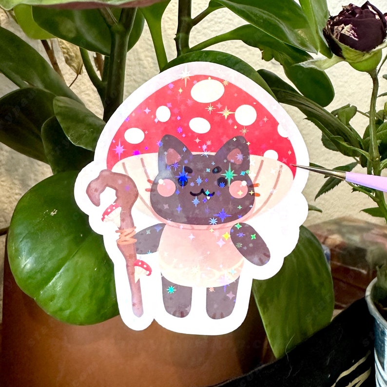 May include: A holographic sticker featuring a cartoon cat character with a mushroom cap hat and a staff. The cat is dark gray with a pink shirt and a white collar. The mushroom cap is red with white spots and star accents.