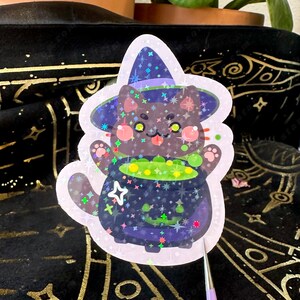 May include: Holographic sticker featuring a cartoon black cat wearing a witch hat. The cat is holding a bubbling cauldron of green potion. The sticker has a pink border and is decorated with stars.