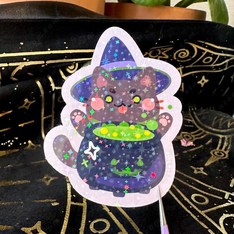 May include: Holographic sticker featuring a cartoon black cat wearing a witch hat. The cat is holding a bubbling cauldron of green potion. The sticker has a pink border and is decorated with stars.