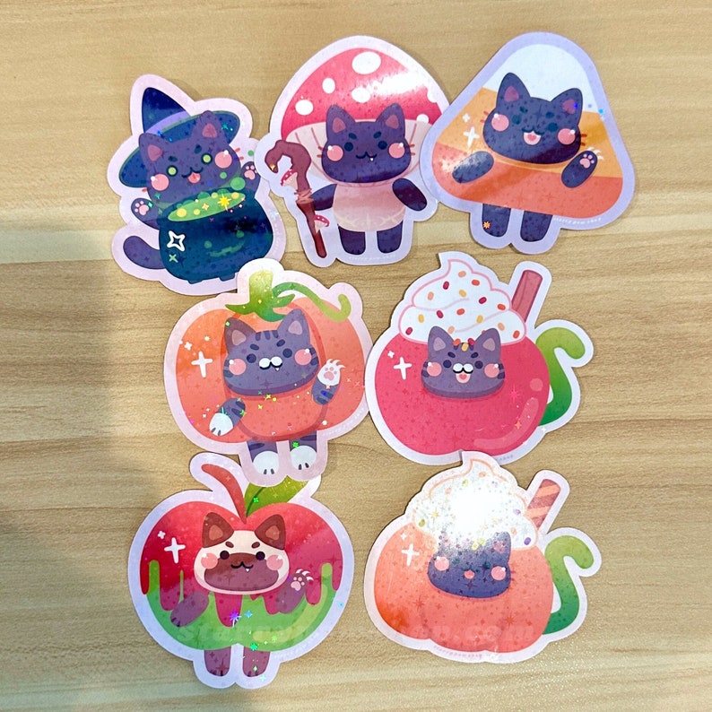 May include: A collection of colorful stickers featuring cartoon cats in Halloween-themed costumes. The stickers include a witch cat, a cat in a mushroom costume, a cat in a candy corn costume, and a cat in a pumpkin costume.