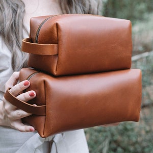 May include: Two brown leather zippered pouches stacked on top of each other. The pouches have a brown leather handle.