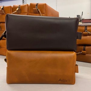 May include: A stack of brown leather travel bags with a zipper closure. The top bag is brown and the bottom bag is tan. The tan bag has the name "Adam" embossed on the front.