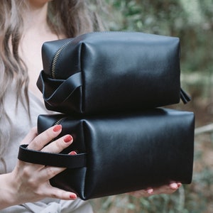 May include: Two black leather zippered pouches stacked on top of each other. The top pouch has a gold zipper and a black leather strap. The bottom pouch has a black leather handle.