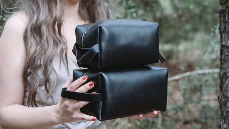May include: Two black leather zippered pouches stacked on top of each other. The top pouch has a gold zipper and a black leather strap. The bottom pouch has a black leather handle.