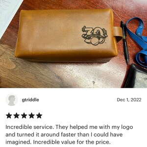 May include: A brown leather toiletry bag with a black leather strap and a black logo that says "70th Anniversary".