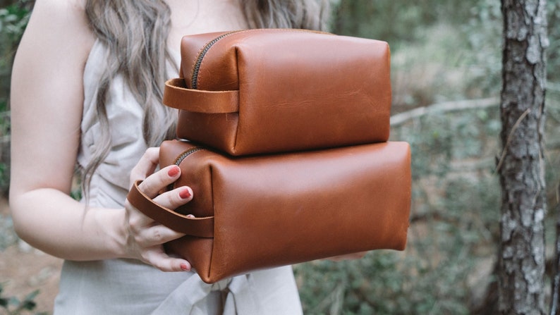 May include: Two brown leather zippered pouches stacked on top of each other. The pouches have a brown leather handle.