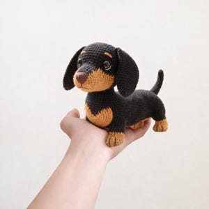 May include: A crocheted dachshund dog in black and tan. The handmade toy has a long body, floppy ears, and a small tail. The dog is held in a person's hand against a white background.