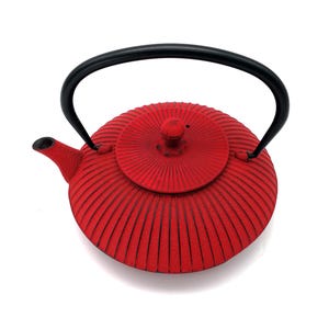 Op de afbeelding: Een ronde, felrode gietijzeren theepot met een textuur van geribbelde patronen die vanuit het midden uitstralen. De pot heeft een bijpassend rood deksel met een kleine knop, een korte tuit en een glad, gebogen zwart metalen handvat.