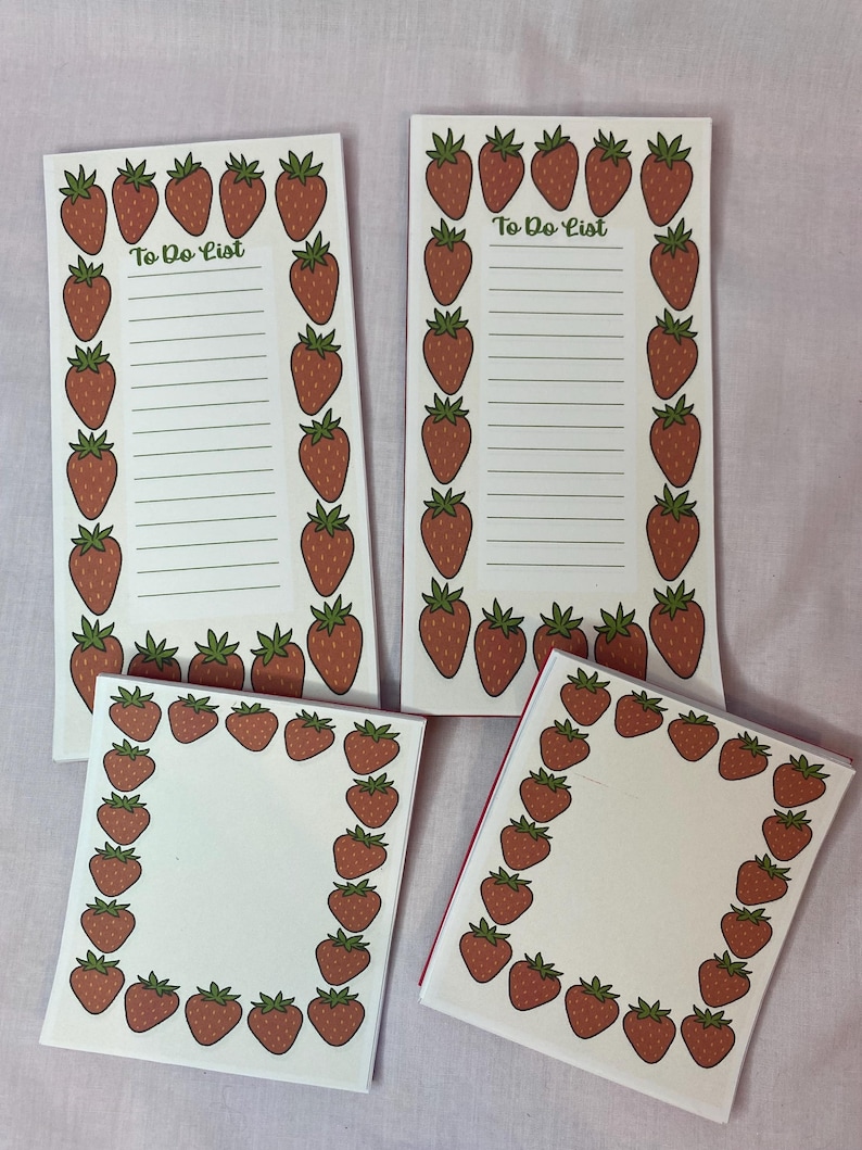 May include: Four white notepads with a strawberry theme. Two rectangular pads have a border of red strawberries and the text "To Do List". The other two pads are square with a strawberry border. The strawberries have green leaves.