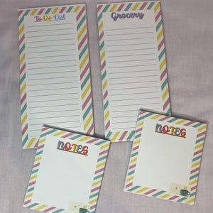 May include: Four notepads with lined pages and colorful diagonal striped borders. Two pads read "To Do List" and "Grocery" in pastel lettering. The other two pads say "Notes" and feature a small sticker with a coffee cup.