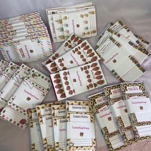 May include: Assortment of notepads with various designs, including strawberries, coffee beans, and leopard print. Some notepads have the words "Grocery List," "Coffee Notes," and "Notes." The brand name "SodaHopShop" is visible on some pads.