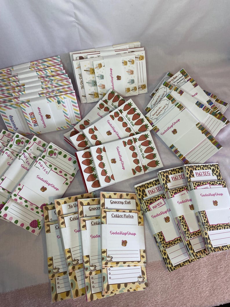 May include: Assortment of notepads with various designs, including strawberries, coffee beans, and leopard print. Some notepads have the words "Grocery List," "Coffee Notes," and "Notes." The brand name "SodaHopShop" is visible on some pads.