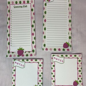 May include: Four notepads with a strawberry theme. Two are rectangular with lined paper, one labeled "Grocery List." The other two are square and say "notes." All have a white background with pink and green polka dots and strawberry illustrations.