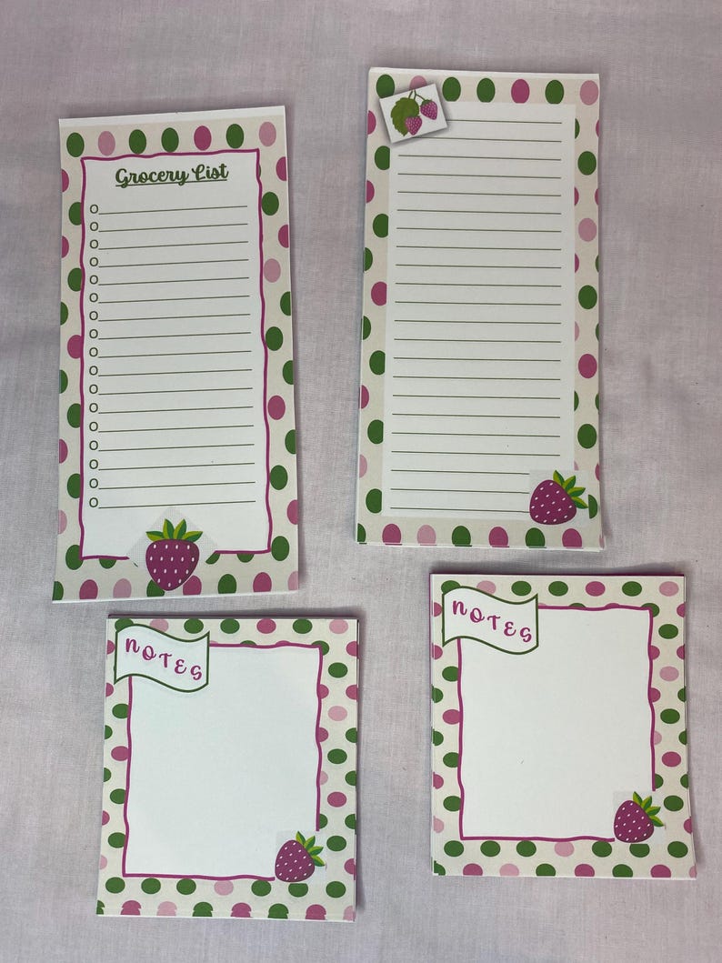 May include: Four notepads with a strawberry theme. Two are rectangular with lined paper, one labeled "Grocery List." The other two are square and say "notes." All have a white background with pink and green polka dots and strawberry illustrations.