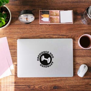 May include: Overhead shot of a wooden desk with various items. A laptop with an "Obedience Dog Approved" sticker is centered. Other items include a plant, coffee, stationery, and a mouse. The desk has a warm, natural wood grain.