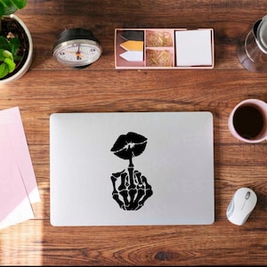 May include: A laptop with a black graphic of a pair of lips and a skeleton hand giving the middle finger. The laptop is on a wooden desk with a coffee cup, mouse, and other office supplies.