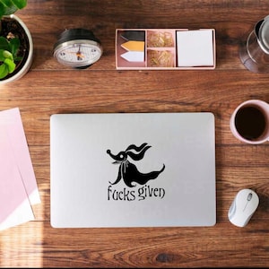 May include: A laptop with a black cartoon dog graphic and the words "fucks given" on a wooden desk. Other items include a plant, a coffee cup, a mouse, and stationery.