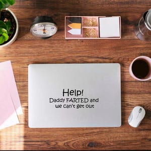 May include: A laptop with the text "Help! Daddy FARTED and we Can't get out" is on a wooden desk. Other items include a plant, a clock, a pink notepad with a pen, a coffee cup, a mouse, and a desk organizer.