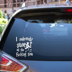 May include: A white vinyl decal applied to the rear window of a car. The decal features the text "I solemnly swear all the f*cking time" in a stylized font, accompanied by a small graphic of a pointed hat and two stars.