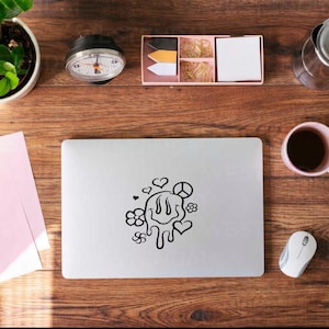 May include: A laptop with a black and white smiley face design, hearts, and flowers is on a wooden desk. Other items include a plant, a clock, a coffee cup, a mouse, and stationery.