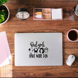 May include: A laptop with the text "Bad girls HAVE MORE FUN" and cartoon villain illustrations is on a wooden desk. A pink mug with coffee, a mouse, and office supplies are also on the desk.