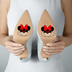 May include: A pair of light-colored pointed-toe high heels featuring a decorative graphic on the sole. The graphic depicts a black mouse silhouette with a prominent red bow and a white ruffled skirt detail centered on the bottom of each shoe.