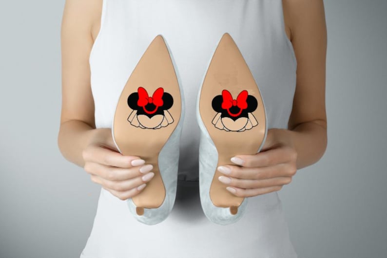 May include: A pair of light-colored pointed-toe high heels featuring a decorative graphic on the sole. The graphic depicts a black mouse silhouette with a prominent red bow and a white ruffled skirt detail centered on the bottom of each shoe.