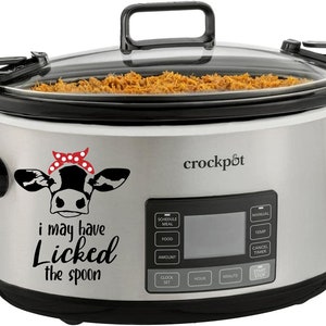 May include: A silver Crock-Pot slow cooker with a black lid and a black handle. The slow cooker has a digital display with buttons for schedule, meal, food, amount, clock set, hour, minute, and start/stop. The slow cooker has a cow with a red and white bandana on its head and the text "i may have licked the spoon".