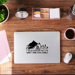 May include: A laptop with a black graphic of a Dementor, the Deathly Hallows symbol, and stick figures. The text reads "THE DEMENTORS DON'T CARE ABOUT YOUR STICK FAMILY." The laptop is on a wooden desk with a coffee cup, mouse, and stationery.