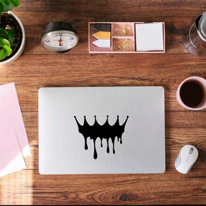 May include: A laptop with a black crown design on the lid sits on a wooden desk. A pink mug with coffee, a white mouse, and a small potted plant are also on the desk. Other items include a clock, a box with office supplies, and a pink notepad.