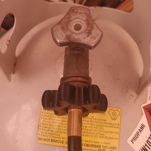 May include: Close-up of a propane tank valve with a black safety handle. The valve has a silver knob with a red warning label that reads "DO NOT REMOVE, DO NOT OBLITERATE THIS LABEL".