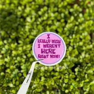 May include: A round sticker with the text "I REALLY WISH I WEREN'T HERE RIGHT NOW!" in purple on a pink background. The sticker is held by tweezers against a backdrop of green foliage. The Lunar Rose Studios logo is at the bottom.