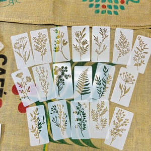 May include: A set of 18 white plastic stencils featuring various floral and leaf designs. The stencils are arranged on a brown burlap background.
