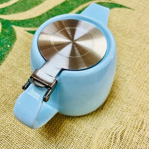 May include: A light blue ceramic teapot with a stainless steel lid and a hinged handle.
