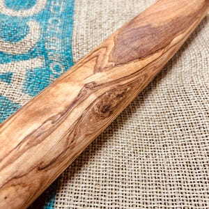 May include: A wooden rolling pin with a natural wood grain pattern. The rolling pin is cylindrical and appears to be made of a light-coloured hardwood, with darker brown streaks and knots. The background is a burlap sack.
