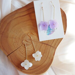 May include: A pair of gold earrings with white and purple pressed flower hydrangea designs. The earrings are on a wooden surface with a card that says "Green Dust Handmade in England".