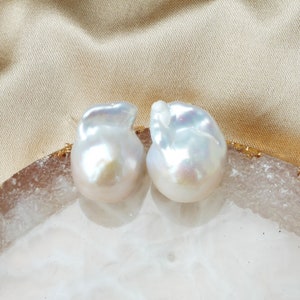Puede incluir: Dos grandes perlas barrocas blancas con un brillo iridiscente. Las perlas se muestran sobre una superficie blanca y dorada.