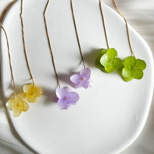 Könnte beinhalten: Drei Paare zarte Ohrringe mit gepressten Blumen. Die Ohrringe sind aus goldenen Ketten gefertigt und zeigen lila, gelbe und grüne Blumen.