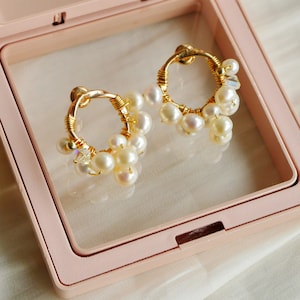 May include: A pair of gold hoop earrings with white pearls and clear crystals hanging from them. The earrings are in a pink jewellery box.