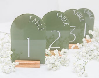 Könnte beinhalten: Vier schildförmige Tischnummern aus grünem Acryl mit abgerundeten Kanten und weißem Text. Die Schilder zeigen "Tisch 1", "Tisch 2", "Tisch 3" und "Tisch 4". Jedes Schild steht auf einem kleinen Holzständer.