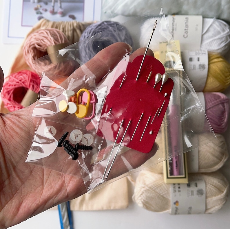 May include: A close-up of a clear plastic bag containing various craft supplies, including needles, pins, buttons, and small black screws. The bag is held in a hand, and there are balls of yarn in the background.