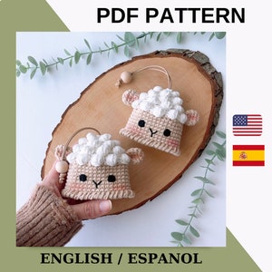 May include: Two crocheted sheep amigurumi toys with brown faces and white wool. The sheep are sitting on a wooden surface with green leaves in the background. The image includes the text "PDF PATTERN" and "ENGLISH / ESPANOL".