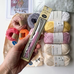 May include: A close-up of a pink crochet hook and a variety of yarn balls in different colors, including pink, yellow, orange, and white. The yarn balls are labeled "Schachenmayr Catania" and are 50 grams each. The crochet hook is labeled "ETIMO Rose 2.20 mm 3/0".