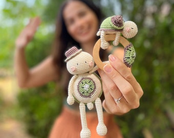 May include: Crocheted baby rattle and toy set featuring a turtle and a character with a hat. The toys are cream, green, and brown, with a wooden ring. The toys are handmade and designed for babies.