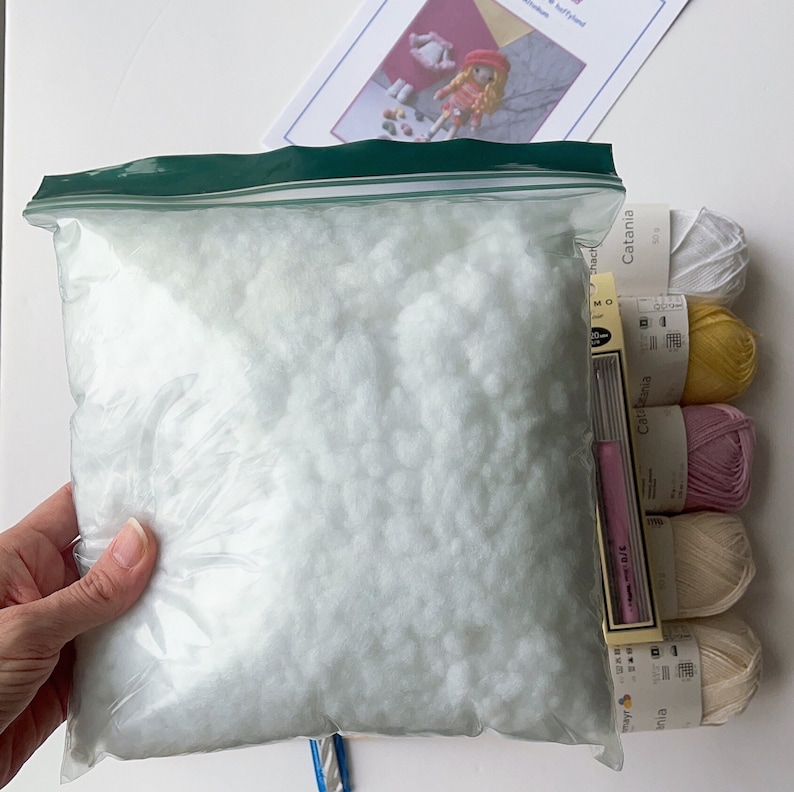 May include: A clear plastic bag filled with white stuffing for a craft project. The bag is next to four balls of yarn in white, yellow, pink, and beige. The yarn is labeled "Catania" and has a size of 50g.