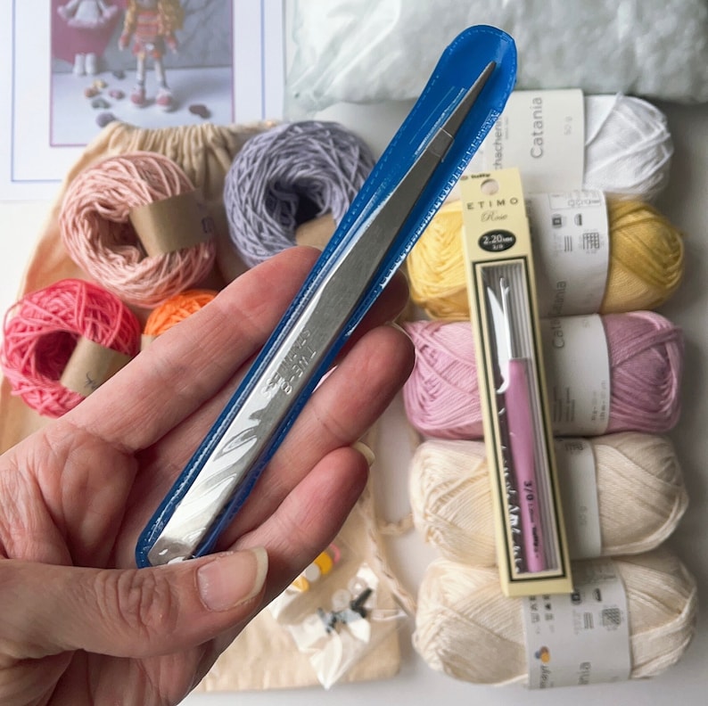 May include: A close-up of a blue handled tweezers with a silver metal tip. The tweezers are held in a hand. The background shows a variety of yarn balls in different colors, including pink, yellow, and white. The yarn balls are labeled "Catania" and "ETIMO".