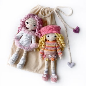 May include: Two crocheted dolls with pink and white clothing. The doll on the left has pink hair and a white dress with pink trim. The doll on the right has blonde hair and a striped dress with a pink bodice.