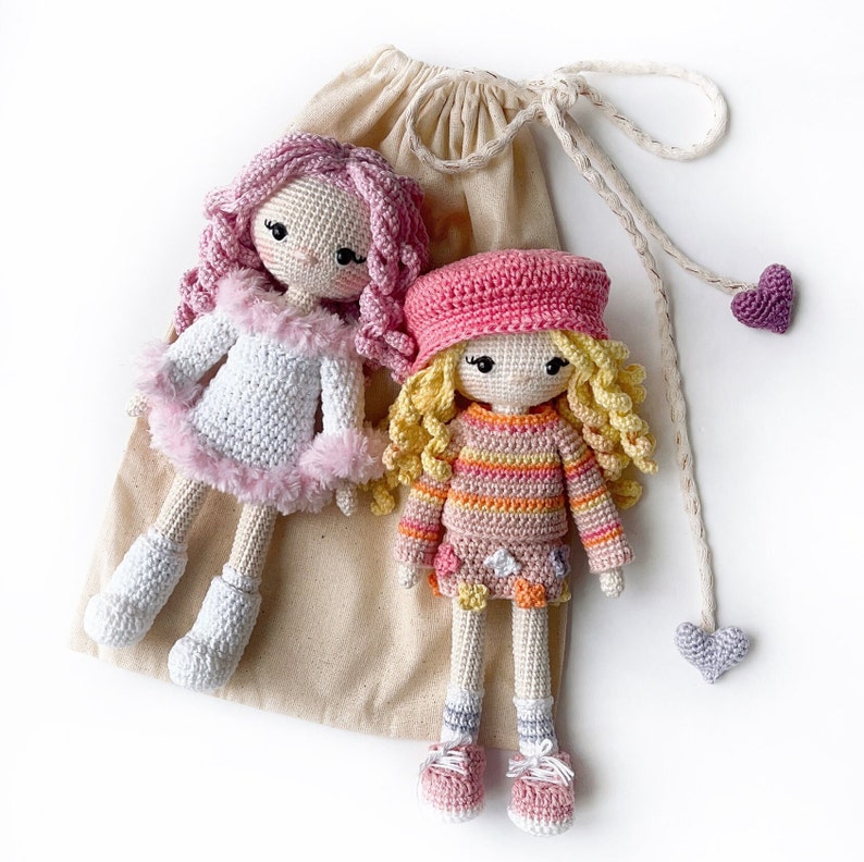 May include: Two crocheted dolls with pink and white clothing. The doll on the left has pink hair and a white dress with pink trim. The doll on the right has blonde hair and a striped dress with a pink bodice.