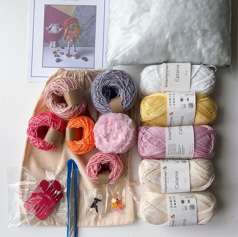 May include: A collection of yarn balls in various colors, including pink, yellow, white, gray, and red. The yarn is labeled "Schachenmayr Catania" and is 50 grams each. There are also small buttons and a needle tool in the image.