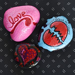 May include: Three hand-painted rocks with heart designs. One pink rock has the word "love" in red script. Another rock features a broken heart with the words "Don't Break My Heart". The third rock has a purple heart design.
